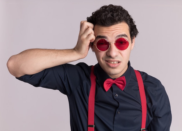 Hombre elegante con pajarita con gafas y tirantes mirando al frente confundido y muy ansioso de pie sobre la pared blanca