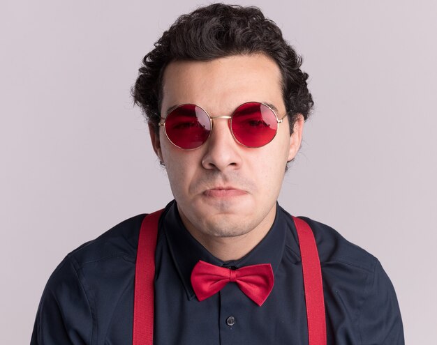 Hombre elegante con pajarita con gafas y tirantes mirando al frente disgustado frunciendo los labios de pie sobre la pared blanca