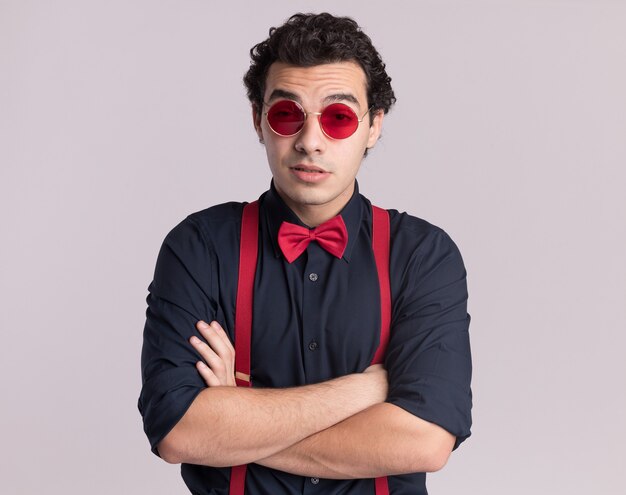 Hombre elegante con pajarita con gafas y tirantes mirando al frente sonriendo confiado con los brazos cruzados de pie sobre la pared blanca