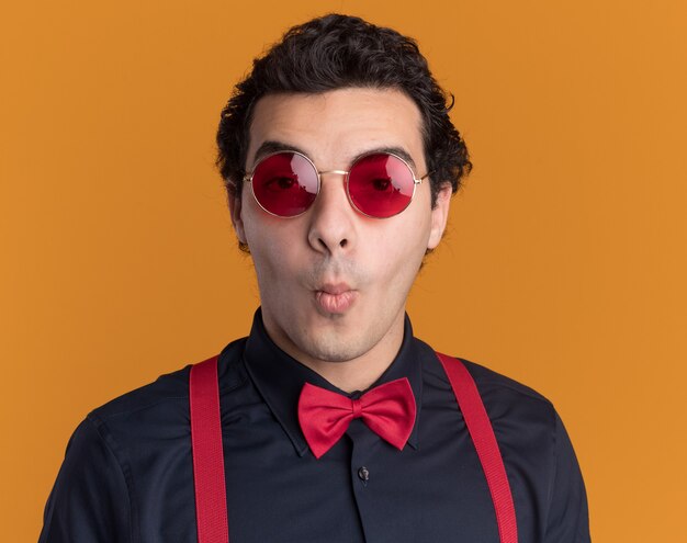 Hombre elegante con pajarita con gafas y tirantes mirando al frente sorprendido parado sobre la pared naranja