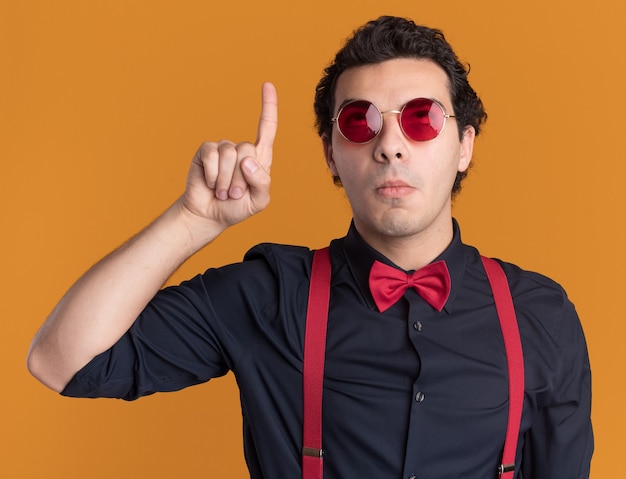 Hombre elegante con pajarita con gafas y tirantes mirando desconcertado apuntando con el dedo índice a algo parado sobre la pared naranja