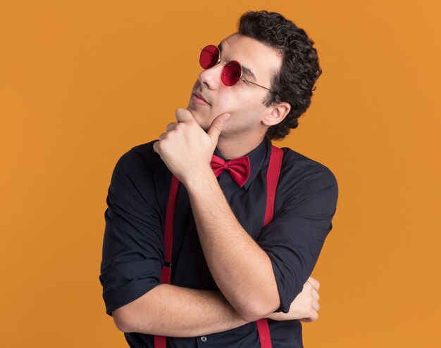 Hombre elegante con pajarita con gafas y tirantes mirando a un lado con la mano en la barbilla pensando de pie sobre la pared naranja