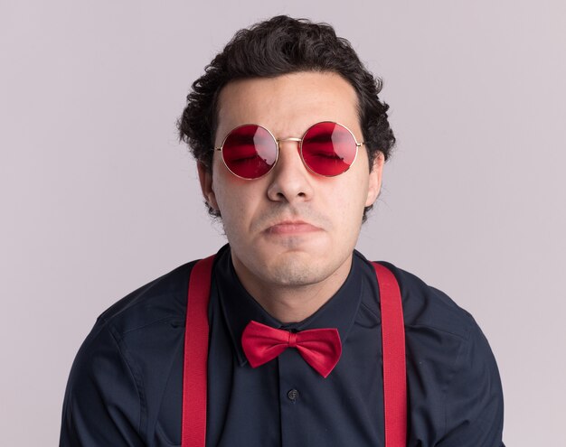 Hombre elegante con pajarita con gafas y tirantes con los ojos cerrados frunciendo los labios de pie sobre la pared blanca