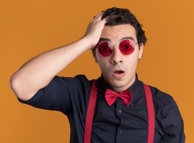 Hombre elegante con pajarita con gafas y tirantes que parece confundido con la mano en la cabeza por error de pie sobre la pared naranja