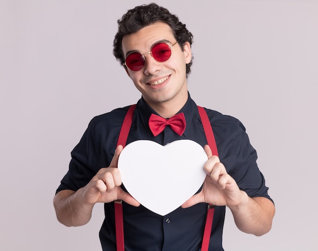 Foto gratuita hombre elegante con pajarita con gafas y tirantes sosteniendo un corazón de cartón mirando al frente sonriendo alegremente de pie sobre la pared blanca