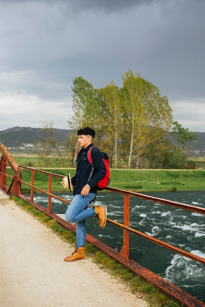 Hombre elegante que sostiene el sombrero que se inclina en la verja del puente sobre el río que fluye