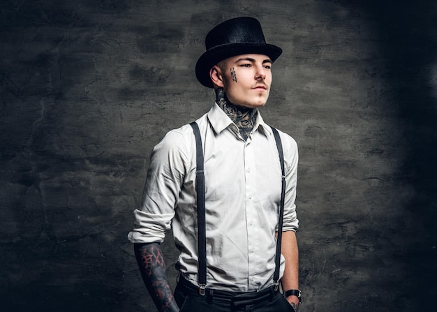 Foto gratuita hombre elegante con tatuajes en los brazos y la cara, vestido con una camisa blanca y un sombrero de copa sobre un fondo gris.