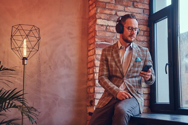 Hombre elegante con traje de franela y gafas escuchando música en una habitación con interior de loft.