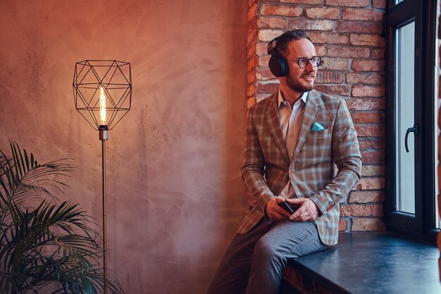 Hombre elegante con traje de franela y gafas escuchando música en una habitación con interior de loft.