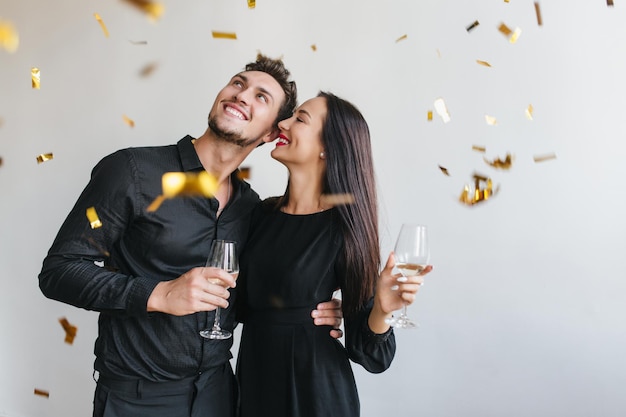 Foto gratuita hombre emocionado con barba negra mirando confeti y abrazando a mujer delgada morena. retrato interior de la feliz pareja amorosa disfrutando de la fiesta con copas de champán.