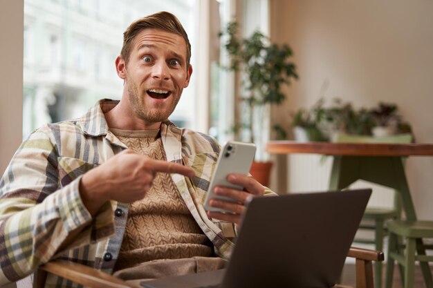 Hombre emocionado en un café sentado con el teléfono y señalando a la pantalla con interés recomendando una aplicación