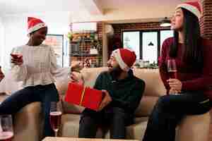 Foto gratuita hombre emocionado con una caja de regalos envuelta de fiesta adivinando el secreto de papá noel mientras celebra la navidad en la oficina compañeros de trabajo alegres con sombreros de navidad intercambiando regalos y disfrutando de las vacaciones de año nuevo