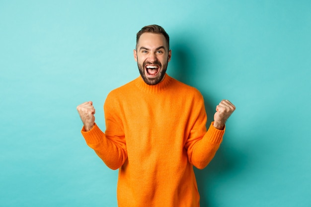 Hombre emocionado celebrando la victoria, regocijándose y haciendo un gesto de bomba de puño, ganando y luciendo satisfecho, diciendo que sí, lograr la meta, de pie sobre una pared turquesa clara.