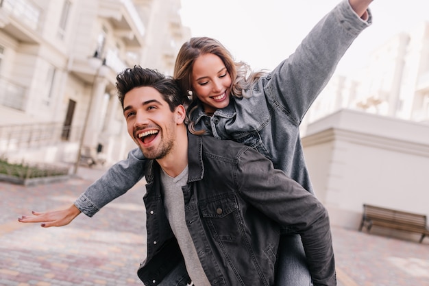 Hombre emocionado en chaqueta de mezclilla negra escalofriante con novia. Retrato al aire libre de la feliz pareja explorando la ciudad.