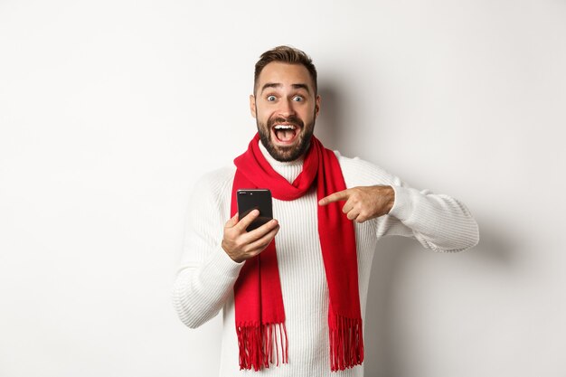 Hombre emocionado hablando de la oferta en el teléfono móvil, apuntando al teléfono inteligente y mirando asombrado, de pie contra el fondo blanco.