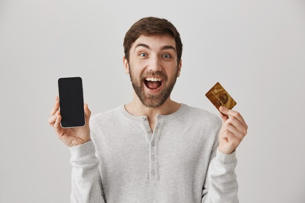Hombre emocionado haciendo un pedido en línea, mostrando la tarjeta de crédito y la pantalla del teléfono móvil