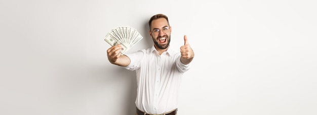 Hombre emocionado mostrando los pulgares hacia arriba y ganando dinero en efectivo de pie sobre fondo blanco.