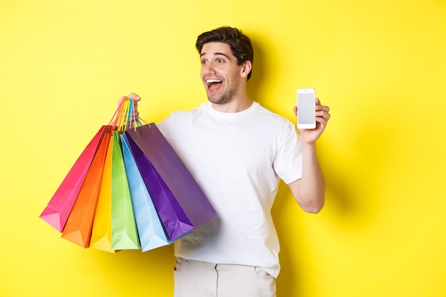Hombre emocionado que muestra la pantalla del teléfono inteligente y bolsas de compras, logra el objetivo de la aplicación, demuestra la aplicación de banca móvil, fondo amarillo.
