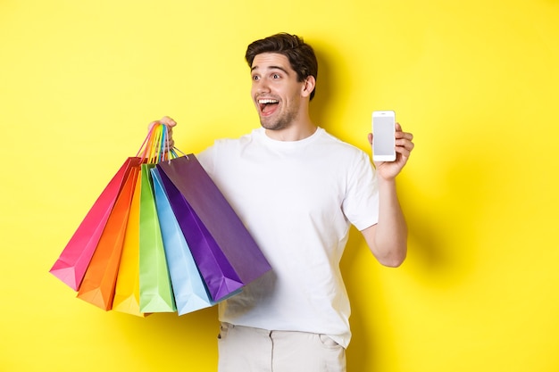 Foto gratuita hombre emocionado que muestra la pantalla del teléfono inteligente y bolsas de compras, logra el objetivo de la aplicación, demuestra la aplicación de banca móvil, fondo amarillo.