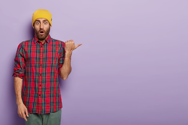 Hombre emocional asustado señala con el pulgar, viste sombrero amarillo y camisa a cuadros, anuncia algo genial