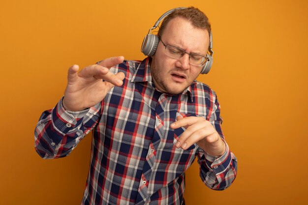 Hombre emocional en gafas y camisa a cuadros con auriculares disfrutando de la música de pie sobre la pared naranja