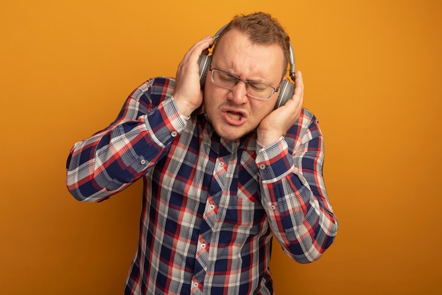 Hombre emocional en gafas y camisa a cuadros con auriculares disfrutando de la música de pie sobre la pared naranja