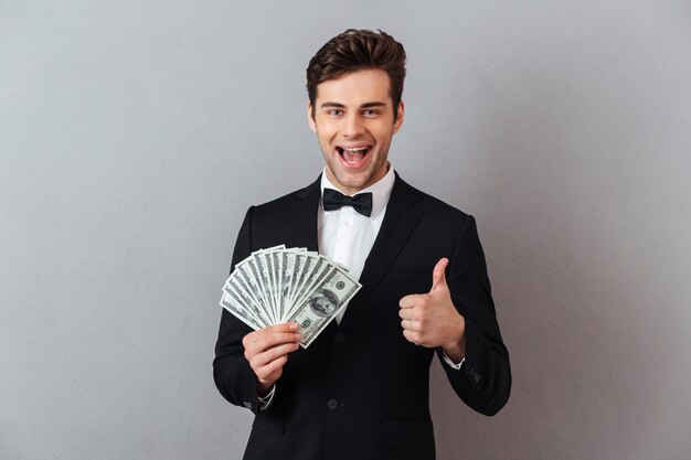 Hombre emocional en traje oficial con dinero mostrando los pulgares para arriba.