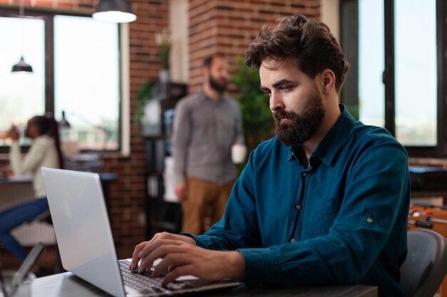 Hombre emprendedor sentado en el escritorio en la oficina de inicio intercambiando ideas mientras trabaja en un proyecto de marketing analizando la facturación de la empresa en una computadora portátil. Diversos empresarios que planean la colaboración empresarial