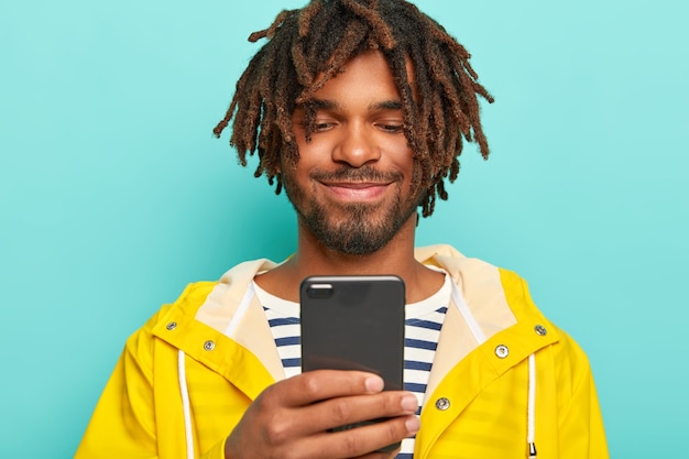 Hombre encantado con piel oscura, tiene rastas, mira positivamente el teléfono inteligente, ve fotos, usa impermeable amarillo