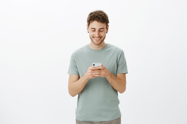 Hombre encantador en aretes, sosteniendo el teléfono inteligente en las manos sobre el pecho y sonriendo ampliamente mientras mira la pantalla