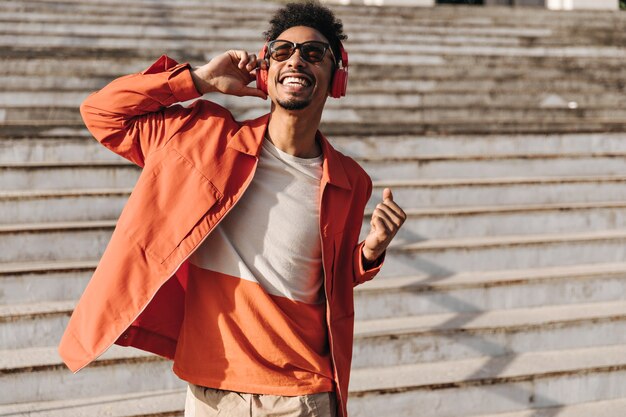 Hombre encantador brunet emocionado con chaqueta naranja, camiseta colorida y gafas de sol canta, sonríe sinceramente y escucha música en auriculares afuera