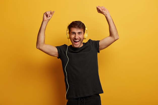 Hombre enérgico guapo levanta los brazos con felicidad, usa auriculares, canta junto con su canción favorita, vestido con una camiseta negra, tiene una expresión de alegría, aislada sobre fondo amarillo