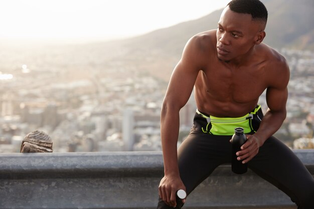 Hombre enérgico con piel oscura, tiene una expresión facial segura de sí mismo a un lado, sostiene una botella con agua, posa en un terreno rocoso, tiene un cuerpo deportivo con músculos. Concepto de fitness, recreación y estilo de vida