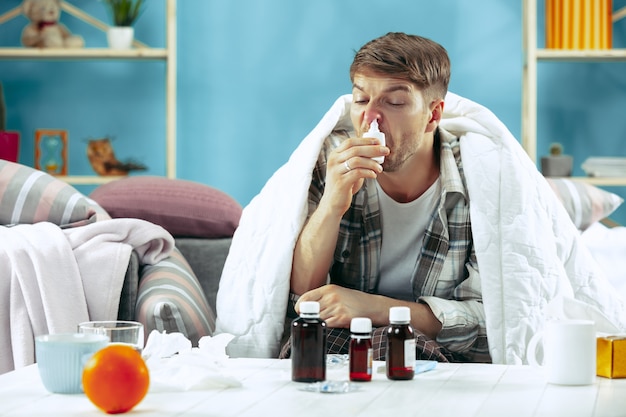 Hombre enfermo barbudo con chimenea sentado en el sofá en casa cubierto con una manta caliente y con spray nasal. La enfermedad, la gripe, el concepto de dolor. Relajación en casa