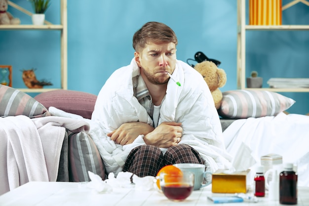 Hombre enfermo barbudo con chimenea sentado en el sofá en casa y midiendo la temperatura corporal. El invierno, la enfermedad, la gripe, el concepto de dolor. Relajación en casa. Conceptos sanitarios.