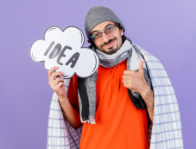 Foto gratuita hombre enfermo caucásico joven complacido con gafas, gorro de invierno y bufanda envuelto en cuadros con burbuja de idea mirando a cámara mostrando el pulgar hacia arriba aislado sobre fondo púrpura