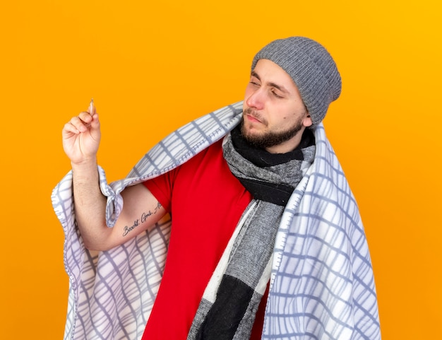 Hombre enfermo caucásico joven confiado con sombrero de invierno y bufanda envuelto en cuadros sostiene y mira el termómetro aislado en la pared naranja con espacio de copia