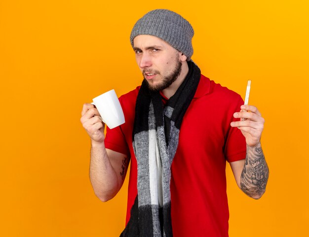 Hombre enfermo caucásico joven disgustado con bufanda y gorro de invierno sostiene taza y termómetro en naranja