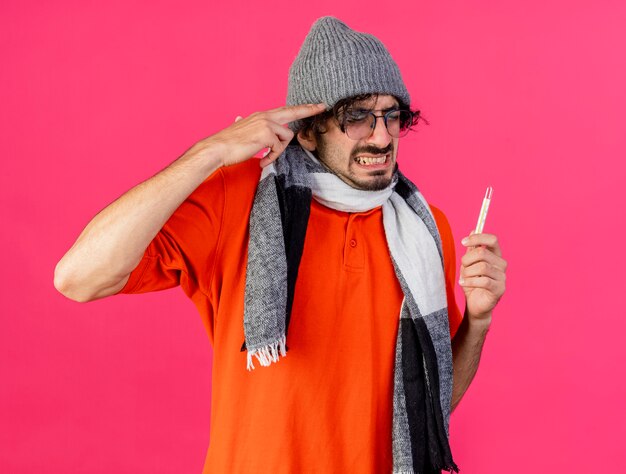 Hombre enfermo caucásico joven estresado con gafas sombrero de invierno y bufanda mirando a cámara sosteniendo termómetro haciendo gesto de suicidio aislado sobre fondo carmesí