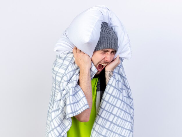 Hombre enfermo caucásico joven molesto con sombrero de invierno envuelto en cuadros sostiene la almohada en la cabeza aislada sobre fondo blanco con espacio de copia
