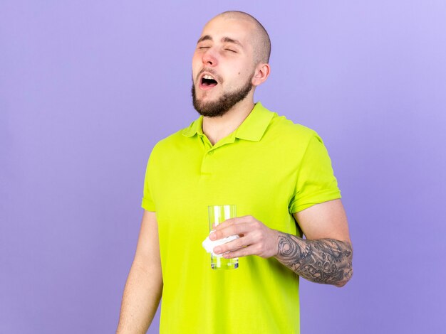 Hombre enfermo caucásico joven pálido quiere estornudar sosteniendo un vaso de agua y tejido aislado en la pared púrpura con espacio de copia
