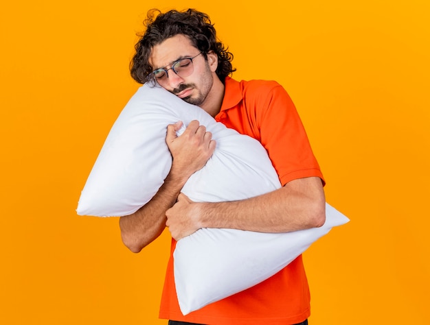 Hombre enfermo caucásico joven soñoliento con gafas abrazando la almohada con los ojos cerrados aislado sobre fondo naranja con espacio de copia