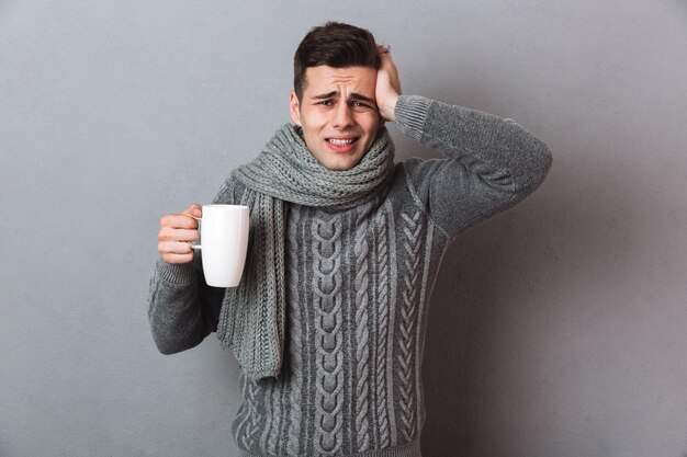 Hombre enfermo confundido en suéter y bufanda que tiene dolor de cabeza mientras sostiene una taza de té y mira