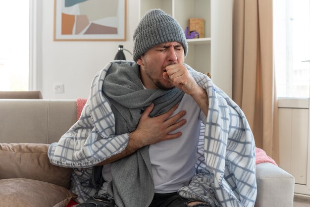 Hombre enfermo dolorido con bufanda alrededor del cuello con gorro de invierno envuelto en cuadros tosiendo manteniendo el puño cerca de la boca sentado en el sofá en la sala de estar