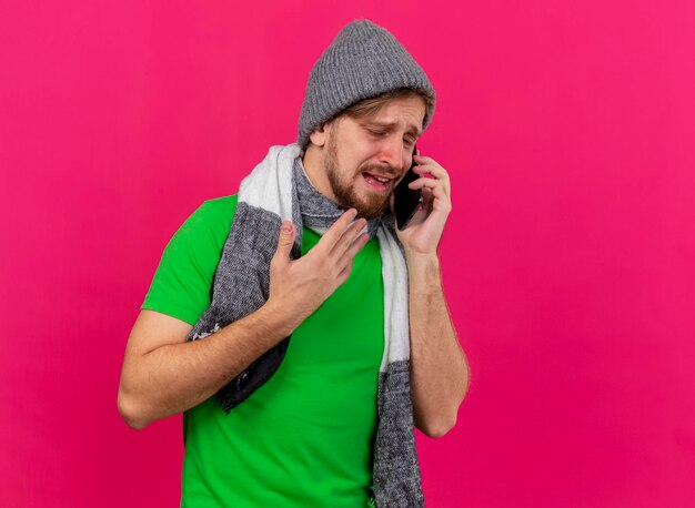 Hombre enfermo eslavo guapo joven disgustado con sombrero de invierno y bufanda hablando por teléfono manteniendo la mano en el aire con los ojos cerrados aislado en la pared rosa con espacio de copia