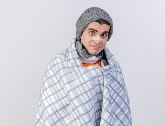Foto gratuita hombre enfermo joven complacido con sombrero de invierno con bufanda envuelta en cuadros con yeso en la nariz aislado en blanco