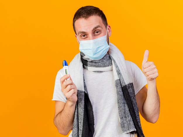 Hombre enfermo joven complacido con sombrero de invierno y máscara médica sosteniendo el termómetro mostrando el pulgar hacia arriba aislado sobre fondo amarillo