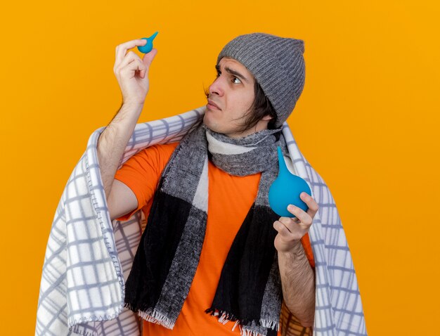 Hombre enfermo joven confundido con sombrero de invierno con bufanda envuelta en cuadros levantando y mirando enema aislado sobre fondo naranja