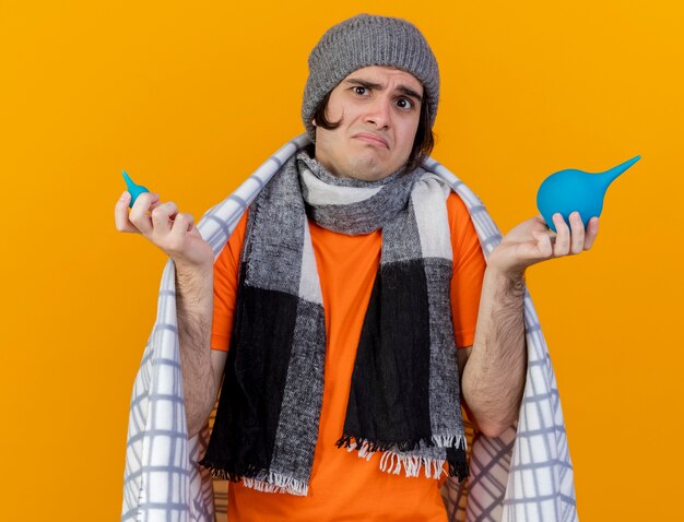 Hombre enfermo joven confundido con sombrero de invierno con bufanda envuelta en cuadros sosteniendo enemas y extendiendo las manos aisladas en naranja