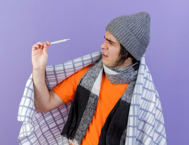 Foto gratuita hombre enfermo joven confundido con sombrero de invierno con bufanda envuelta en cuadros sosteniendo y mirando el termómetro aislado en púrpura